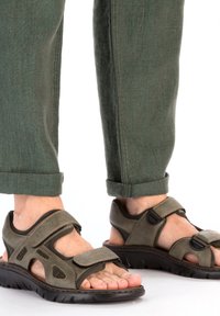 Des sandales en cuir marron avec des brides réglables, des sections en mesh respirant et une semelle en caoutchouc noir flexible. Portées avec un pantalon vert retroussé.