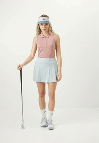 Fiatal nő golf öltözékben, golfütővel a kezében, rózsaszín, ujjatlan pólót, világoskék szoknyát, fehér napernyőt és sportcipőt visel.