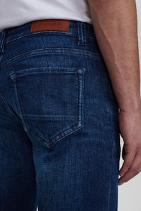 Temno modri jeans iz denima z zadnjim žepom z vidnimi šivi in rjavim usnjenim oznako na pasu, ki prikazuje "GABBA."