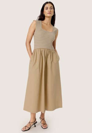 Robe beige avec un col carré côtelé et un haut ajusté, s'évasant en une jupe ample arrivant jusqu'aux chevilles. Comprend des poches latérales et est portée avec des sandales noires.