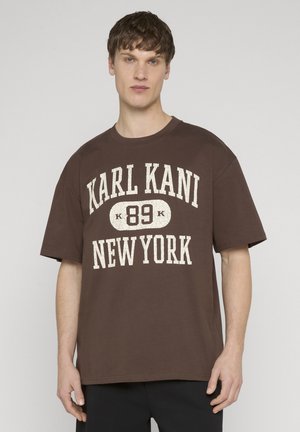 Jaunas vyras vilki rudą, per didelį marškinėlį su užrašu „KARL KANI 89 NEW YORK“ baltais rašmenimis priekyje, priešais paprastą šviesų foną.