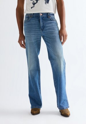 Hombre vistiendo jeans azul claro de pierna recta, camiseta blanca con diseño gráfico y botas de ante marrón sobre fondo blanco.