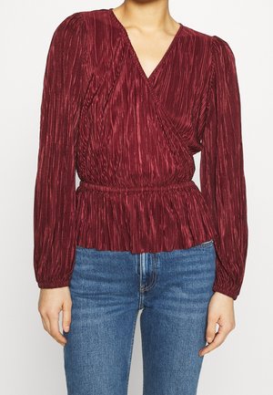 Donna che indossa una camicetta plissettata a manica lunga di colore rosso scuro con scollo a V e vita elasticizzata, abbinata a jeans blu.