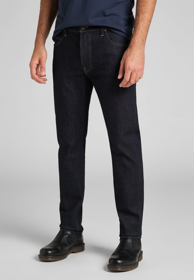 Jeans de mezclilla azul oscuro con un corte ajustado, que cuentan con costuras naranjas, cierre de botón y bolsillos delanteros. Llevados con botas de cuero negras.