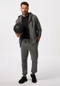Sudadera atlética gris con bolsillos frontales, combinada con joggers a juego y zapatillas deportivas negras, sosteniendo una pelota de medicina negra.