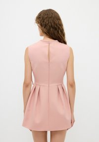 Robe sans manches rose avec un corsage ajusté, une jupe plissée et un détail en trou de serrure au dos. Fabriquée en tissu lisse, elle arrive au-dessus du genou.