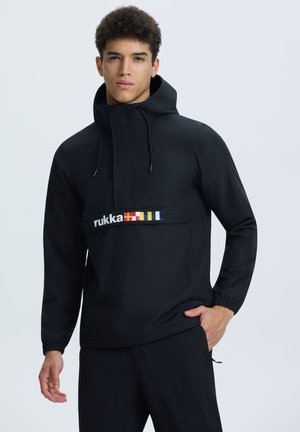 Nuori mies, joka on pukeutunut mustaan huppariin, jossa on "rukka"-logo ja värikkäitä lippuja rinnassa, poseeraa käsi taskussa.