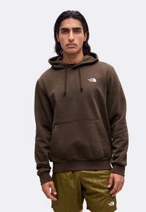 Uomo con capelli neri fino alle spalle, che indossa una felpa con cappuccio marrone The North Face e pantaloncini verdi The North Face, in piedi davanti a uno sfondo semplice.