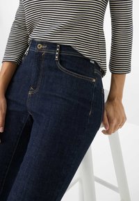 Jean en denim bleu foncé avec un design à cinq poches, des coutures dorées et une fermeture à bouton. Associé à un haut à rayures noir et blanc.