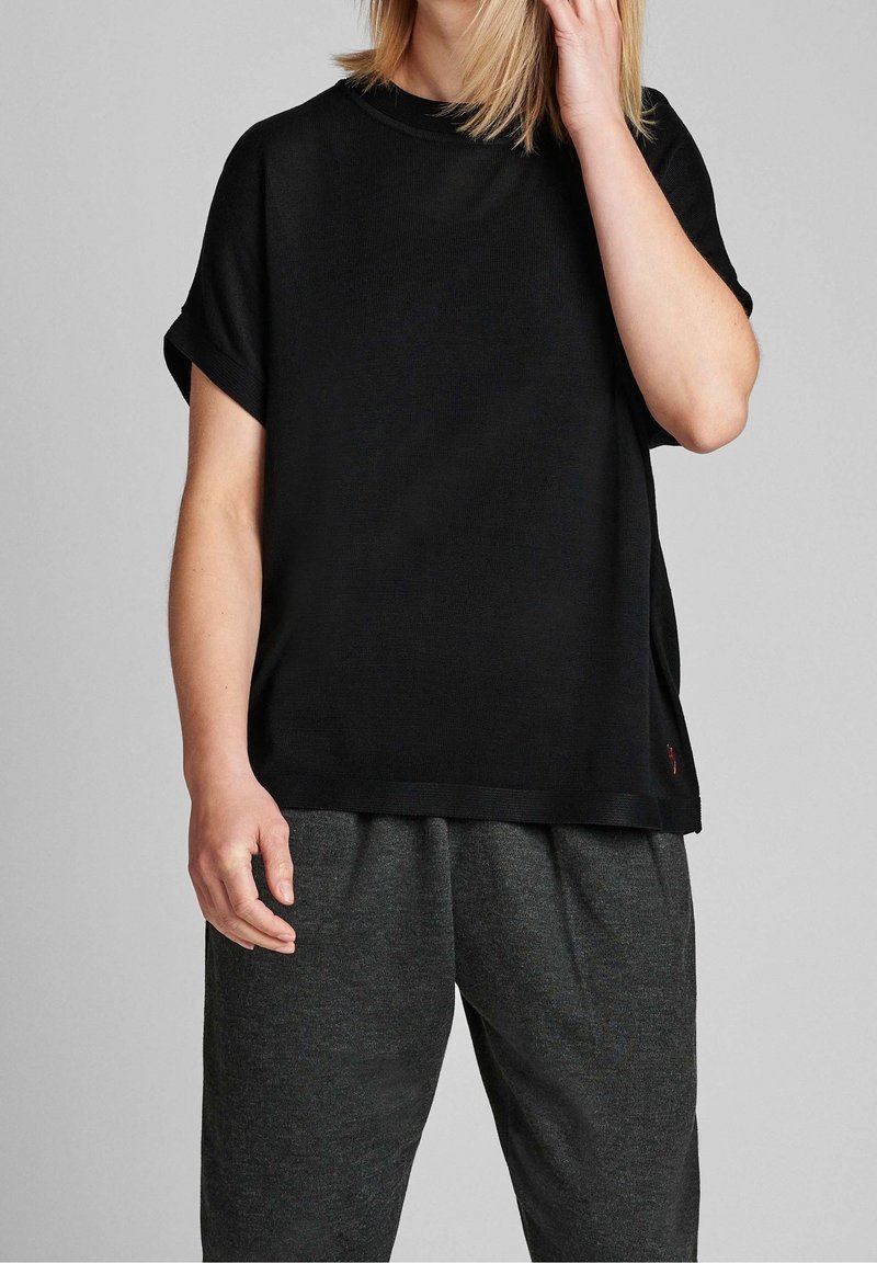 Schwarzes Kurzarmhemd mit Rundhalsausschnitt, aus einem glatten Stoff gefertigt, mit umgeschlagenen Ärmeln und einem lockeren Schnitt. Kombiniert mit dunkelgrauen Hosen.