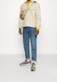 Chaqueta beige clara con cremallera sobre una camiseta amarilla, combinada con vaqueros azules y zapatillas verdes/morado. Un bolso cruzado color canela se lleva al pecho.