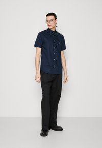 Young man with slicked-back hair wearing clear glasses, navy short-sleeve button-up shirt, black pants, and black shoes, standing neutral pose.