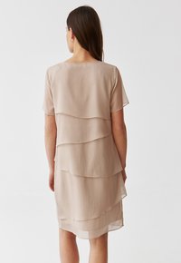 Beige kerroksellinen mekko, joka on valmistettu valuvaista, laskostettua kankasta. Mekossa on lyhyet hihat ja pyöreä pääntie sekä kolme erillistä päällekkäistä tasoa.