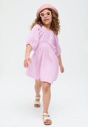 Junges Mädchen in einem pinken Kleid, passendem Hut, sonnenbrille in Blumenform und weißen Sandalen, das selbstbewusst vor einem weißen Hintergrund geht.