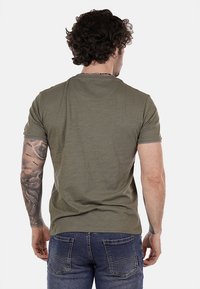 T-shirt verde oliva a maniche corte con scollatura rotonda e finitura testurizzata, abbinato a jeans in denim blu, che mostra un braccio tatuato.