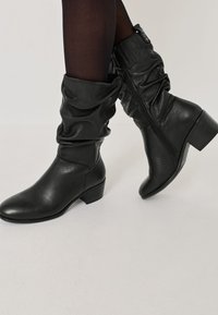 Cizme din piele negre, până la jumătatea gambei, cu un aspect lejer și fermoar pe lateral, purtate cu ciorapi negri subțiri pe un fundal deschis.