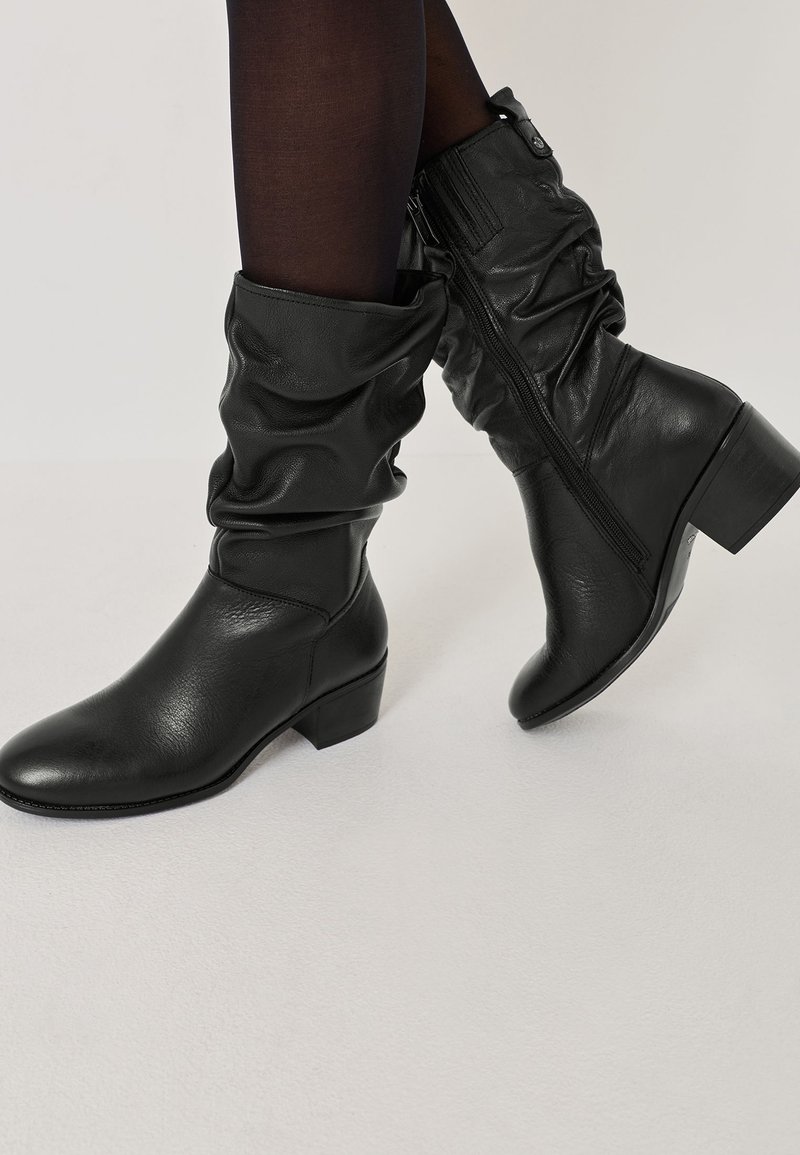 Cizme din piele negre, până la jumătatea gambei, cu un aspect lejer și fermoar pe lateral, purtate cu ciorapi negri subțiri pe un fundal deschis.