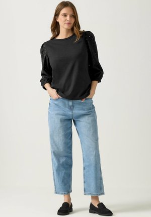 Frau steht mit den Händen in den Taschen, trägt ein schwarzes Oberteil mit Puffärmeln, hellblaue Jeans mit weitem Bein und schwarze Loafers vor einem einfachen Hintergrund.