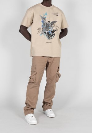 T-shirt graphique beige avec un design imprimé et des accents bleus, associé à un pantalon cargo marron clair doté de plusieurs poches et d'une coupe décontractée.
