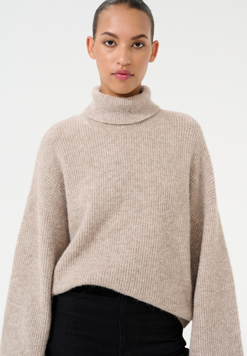 Maglione a collo alto beige a costine con una vestibilità rilassata e maniche ampie, realizzato in un morbido materiale a maglia. Sotto il maglione sono indossati dei jeans neri.