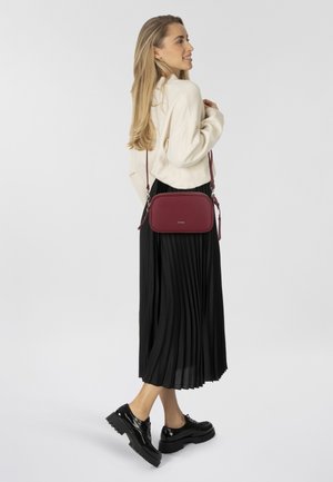 Borsa a tracolla in pelle bordeaux con una texture liscia, forma rettangolare e tracolla regolabile. Una gonna nera plissettata e scarpe con plateau nere completano il look.