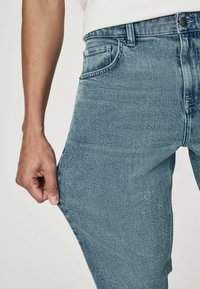 Jeans din denim de un albastru deschis, cu un design cu picior drept, având un buzunar frontal, cusături vizibile și o mână care prinde talia.