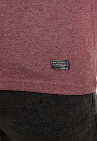 T-shirt di cotone bordeaux con un'etichetta nera rettangolare che riporta la scritta "AUTHENTIC BERLIN DESIGN IVY CORE" vicino all'orlo. Texture morbida, design casual.