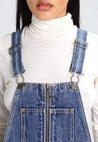 Denim overalls with adjustable blue straps, a silver zipper front, and two front pockets. The fabric appears textured with orange stitching.