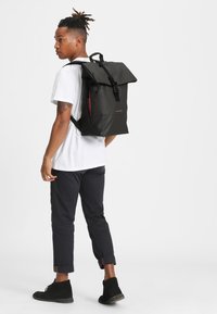 Black roll-top backpack with adjustable straps, a front zipper pocket, and a smooth matte finish. Features a logo on the front panel.