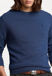 Personne portant un pull en maille bleu avec un petit logo de cheval sur la poitrine et une ceinture en cuir marron avec une boucle argentée.