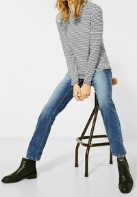 Černé kotníkové kožené boty, modré úzké džíny a dlouhý pruhovaný rukáv. Osoba sedí na hnědé kovové stoličce.