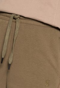 Pantalones de chándal verde oliva con cintura ajustable, que presentan un acabado texturizado y un pequeño logo bordado en amarillo en el lado inferior.
