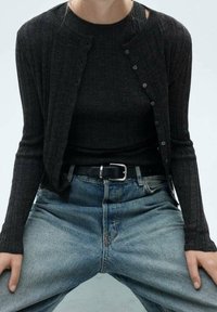 Cardigan noir côtelé avec col rond, fermeture boutonnée, associé à un haut noir et un jean denim bleu, accentué par une ceinture noire.