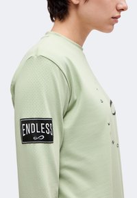 Personne portant une chemise à manches longues vert clair en tissu perforé avec un patch noir sur la manche indiquant "ENDLESS" avec le symbole de l'infini.