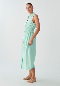 Mėtų žalios spalvos suknelė be rankovių, su mygtukų uždarymu priekyje, elastinga juosmeniu ir iki kelių ilgio. Lygus audinys su laisvu siluetu.