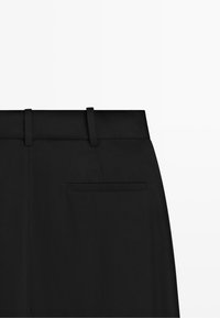 Close-up van de bovenrug van een zwarte broek met riemlussen en een enkele bieszak op een effen achtergrond.