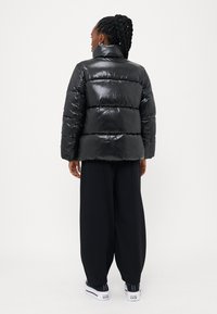 Sort pufferjakke med en blank overflade, cropped design, vandrette quiltede sektioner, høj krave og brede ærmer.