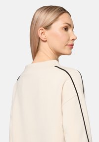 Jasnobeżowa bluza o swobodnym kroju, z czarnymi paskami na ramionach i okrągłym dekoltem. Miękka w dotyku.