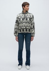 Bijeli i crni pulover od flisa s uzorkom, s polu-zip zatvaračem, s geometrijskim dizajnom. Kombiniran s tamnoplavim širokim trapericama i bijelim tenisicama.