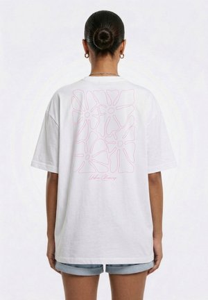 Kvinde med hår i knold iført hvid oversize T-shirt med pink abstrakte blomsterdesign og tekst bagpå, kombineret med denimshorts.