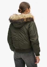 Olijfgroene bomberjack met een nep bont gevoerde capuchon, geribbelde manchetten en zoom; heeft een gewatteerde textuur aan de buitenkant.
