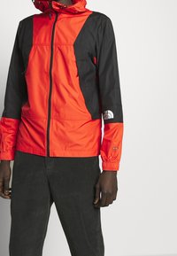 Červeno-černá větruodolná bunda na zip, s elastickými manžetami a logem. Vyrobena z lehkého materiálu, s kapucí a designem s blokovými barvami.