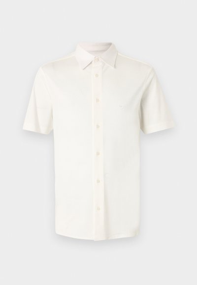 Λευκό πουκάμισο με κοντά μανίκια και κουμπιά, με μυτερό γιακά και διακριτικό λογότυπο στο αριστερό στήθος, παρουσιασμένο σε απλό ανοιχτό γκρι φόντο.