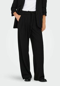 Donna che indossa pantaloni neri a gamba larga con una cintura annodata e un blazer nero sopra una maglietta bianca, in piedi con una mano in tasca.