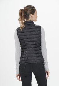 Chaleco de plumas negro con un alto cuello, que presenta un patrón acolchado y un diseño ligero, usado sobre una camiseta de manga larga gris y jeans negros.