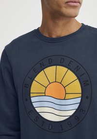 Tamsiai mėlynas džemperis su apvaliu grafinėmis, vaizduojančiomis saulę ir bangas. Tekstas „BLEND DENIM“ ir „ESTD 1993“ juoda spalva. Minkšta medžiagos tekstūra.