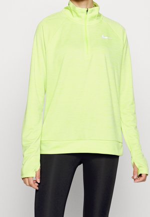 Persoon draagt een neongele lange mouwen sporttop met een kwart rits en duimgaten, gecombineerd met zwarte legging.