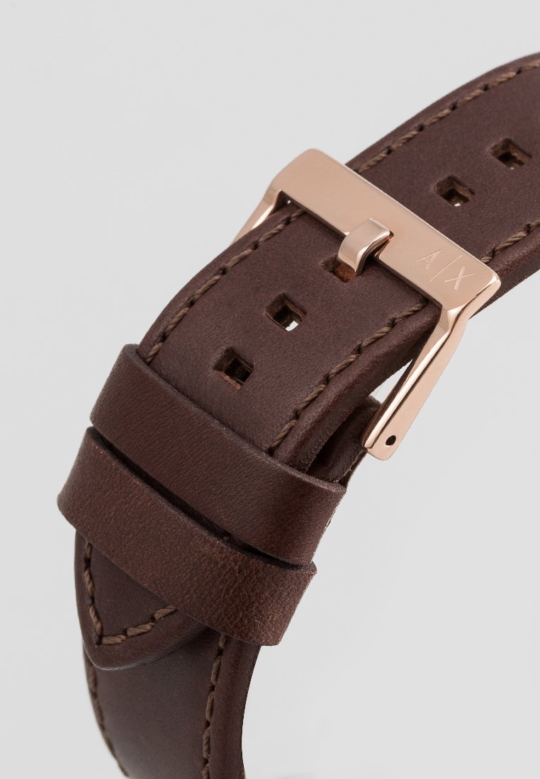 Brązowy skórzany pasek do zegarka o gładkiej fakturze, z różowo-złotą metalową klamrą oraz kilkoma otworami do regulacji długości.