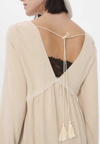 Beige jurk met een diepe V-rug met strikjes, kantafwerking en twee franjes. Zwarte kant bralette zichtbaar onder de jurk. Gladde stoftextuur.