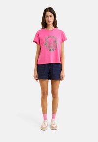Camiseta de algodón rosa con mangas cortas, con un gráfico verde que dice "VACACIONES EN", combinada con pantalones cortos de mezclilla oscura y zapatillas blancas.